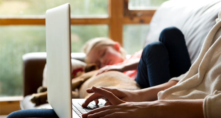 mother working on laptop while child sleeps on couch