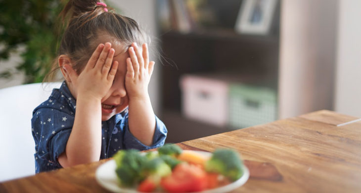 child covering her eyes in front of plate of veggies