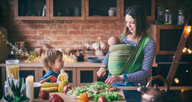 Happy young family, beautiful mother with two children, adorable preschool boy and baby in sling cooking together in a sunny kitchen.